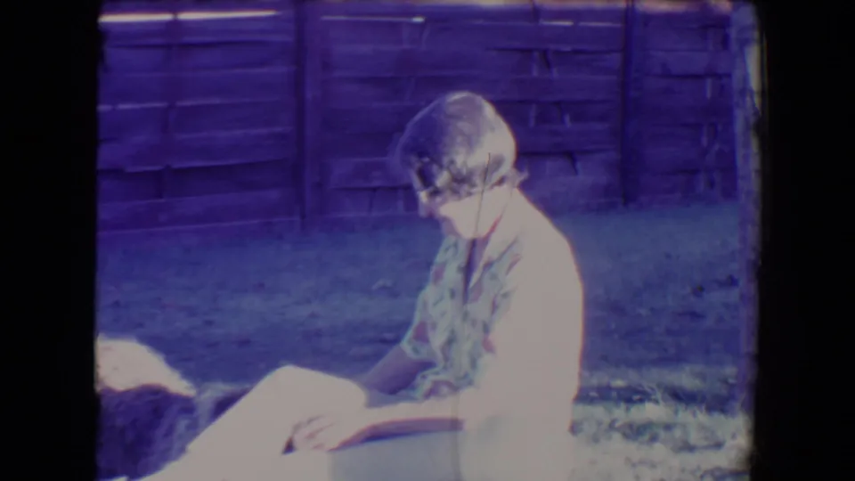 1967 Tucson 8mm frame of a seated woman in bright sunlight with the dog partly visible at the edge of the grassy yard scene.