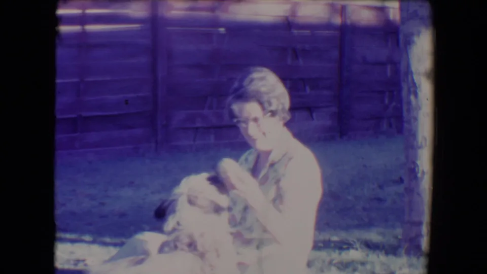 1967 Tucson home-movie still of a seated woman interacting with a shaggy dog on grass, with aged color shift and fence behind.
