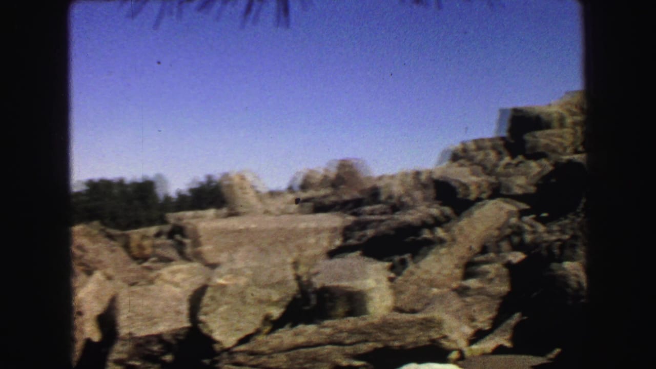 Rocky boulder field with distant trees and blue sky, partially framed by dark film edges.