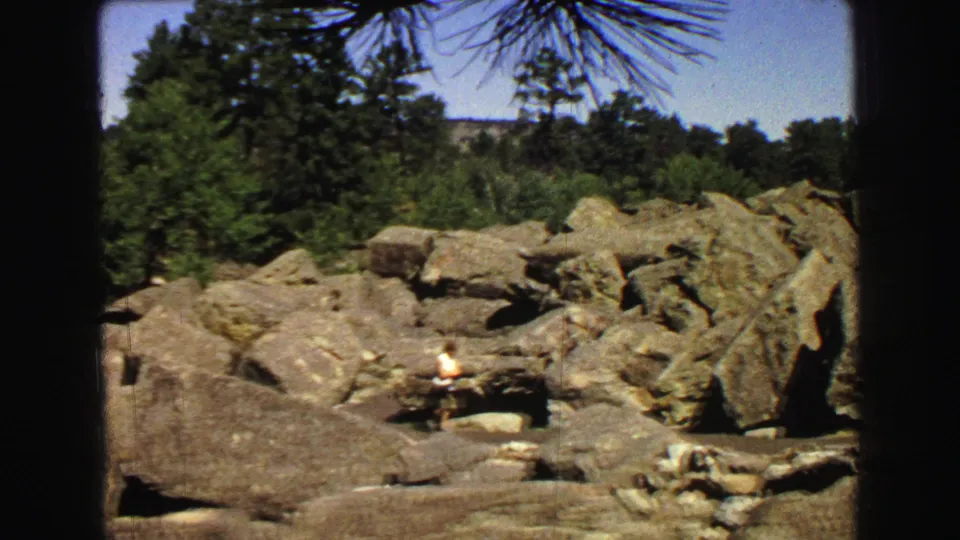1967 Yellowstone view of a distant person in light clothing among massive boulders, with green trees and pine branches framing the scene.