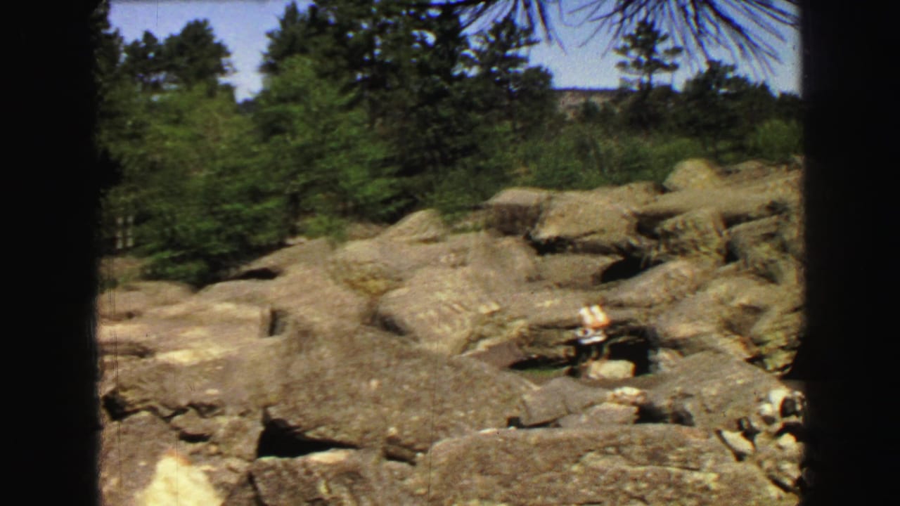 Rocky pine-covered slope with a small person standing on the ledges.