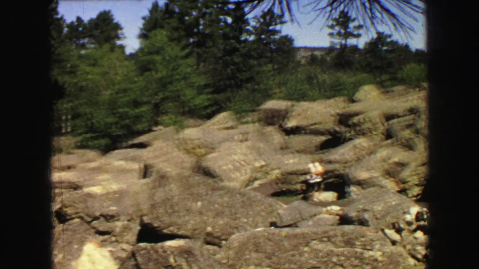 1967 8mm travel frame showing pale rock ledges, pine trees, and a distant visitor in light clothing.