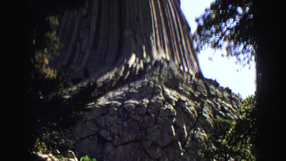 Handheld 8mm view of Devils Tower's vertical columns and rough lower rock face framed by trees.