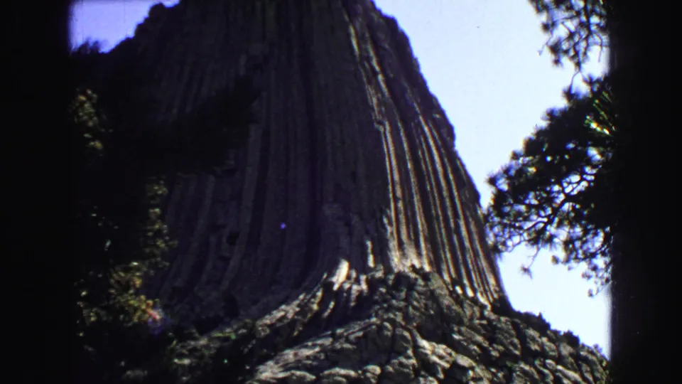 1967 8mm frame of Devil's Tower rising behind trees, with vertical rock columns and shadowed foreground foliage.