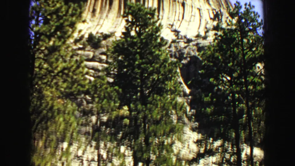 Visitor-shot 1967 8mm frame showing Devil's Tower above trees, with a rocky base and sunlit vertical columns.