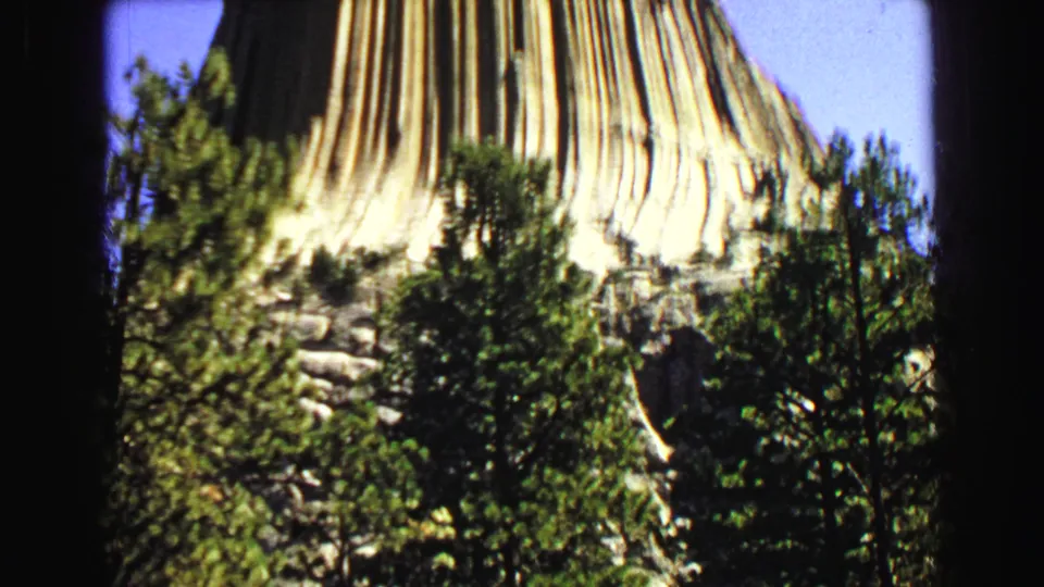 1967 8mm frame showing the lower rock columns of Devils Tower with pine trees at the base and blue sky above.