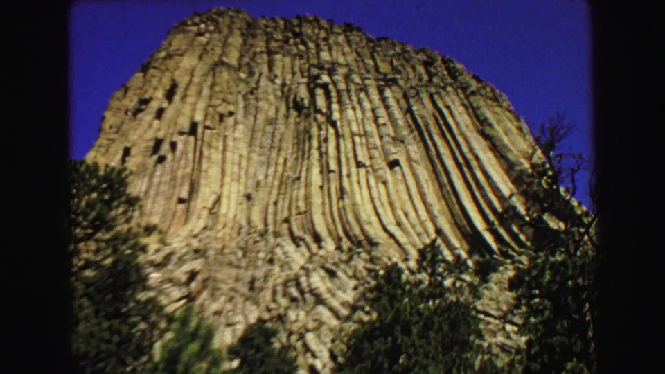 1967 8mm frame showing a broad sunlit face of Devils Tower, with vertical columns, trees at the base, and clear sky.