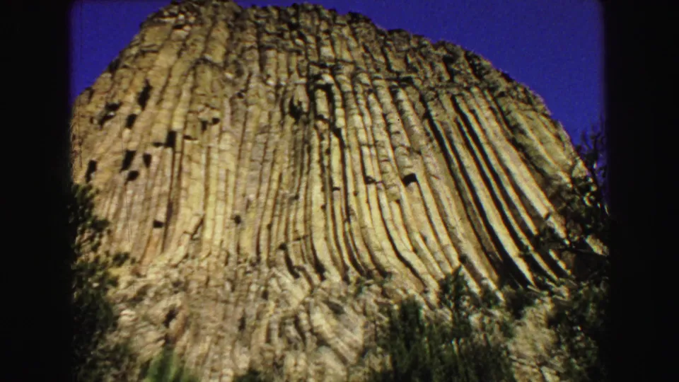 1967 8mm frame showing a low-angle view of Devils Tower's vertical rock columns against blue sky, with trees near the base.