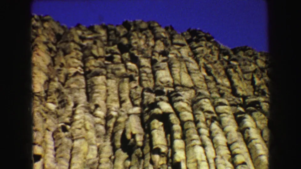 Tight 1967 8mm view of Devils Tower's upper rock face, with fractured vertical columns silhouetted against saturated blue sky.
