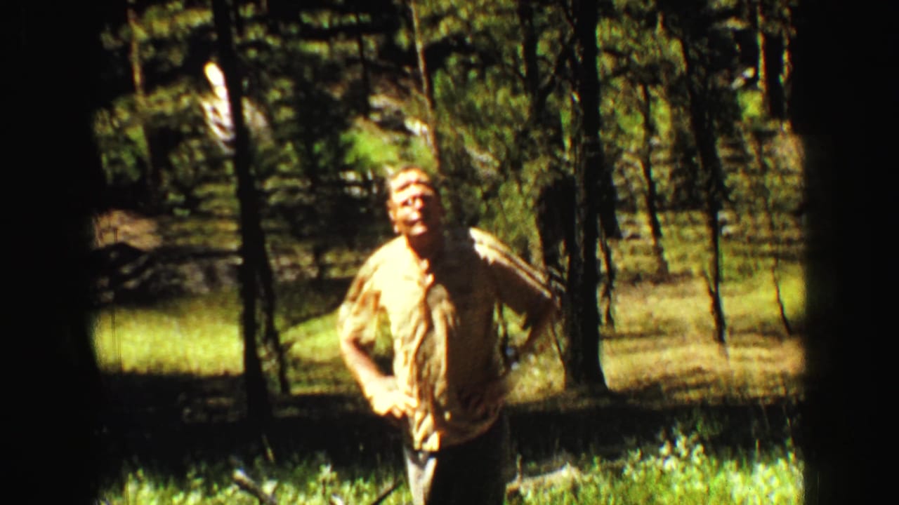 Man in a yellow shirt waving while standing among trees.