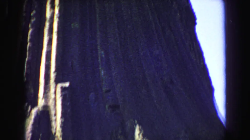 1967 Yellowstone home-movie frame looking up at the textured vertical cliff, with dark exposure and pale sky at the side.