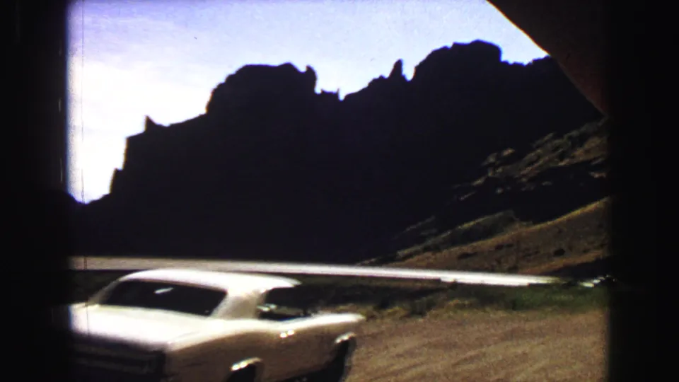 White mid-century car driving through Yellowstone mountain terrain in a 1967 8mm home-movie frame.