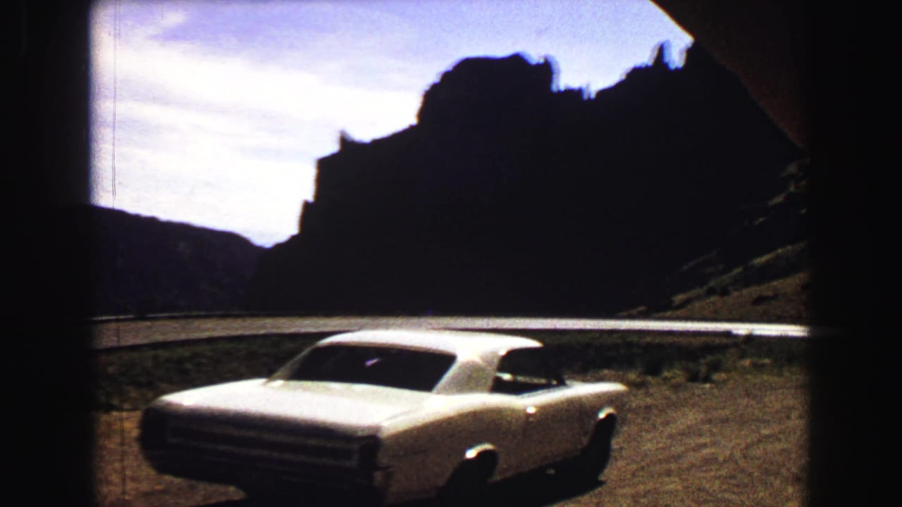White coupe parked on a dirt overlook below dark hills and a bright sky.