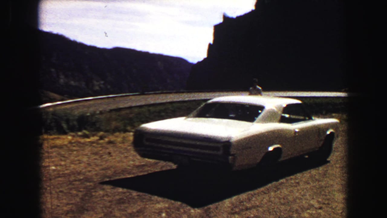 White coupe seen from the rear quarter near a curving road and rocky hillside.