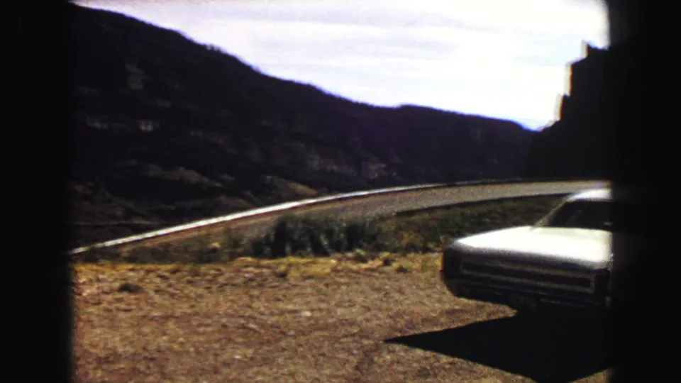 1967 8mm frame with the front of a white coupe partly cropped at a Yellowstone-area overlook, road and hills in the background.