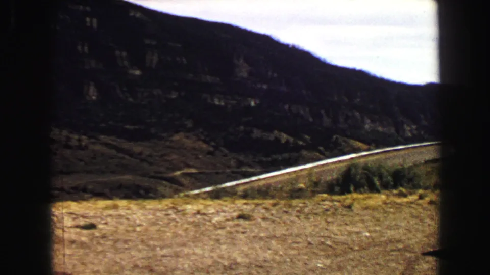 1967 Yellowstone-area 8mm landscape view of a winding road, guardrail, rocky hills, and the edge of the white car at right.