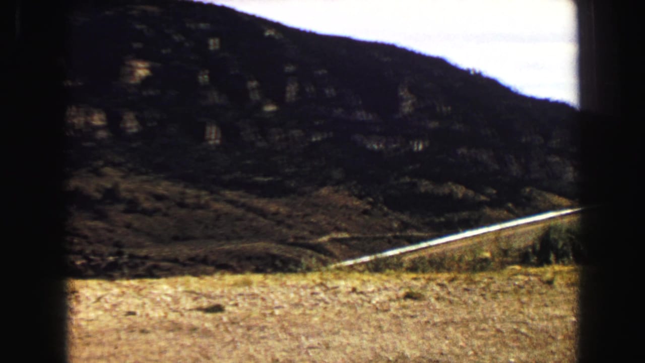 Rocky mountain slope with dry grass and a pale road seen from a moving vehicle.