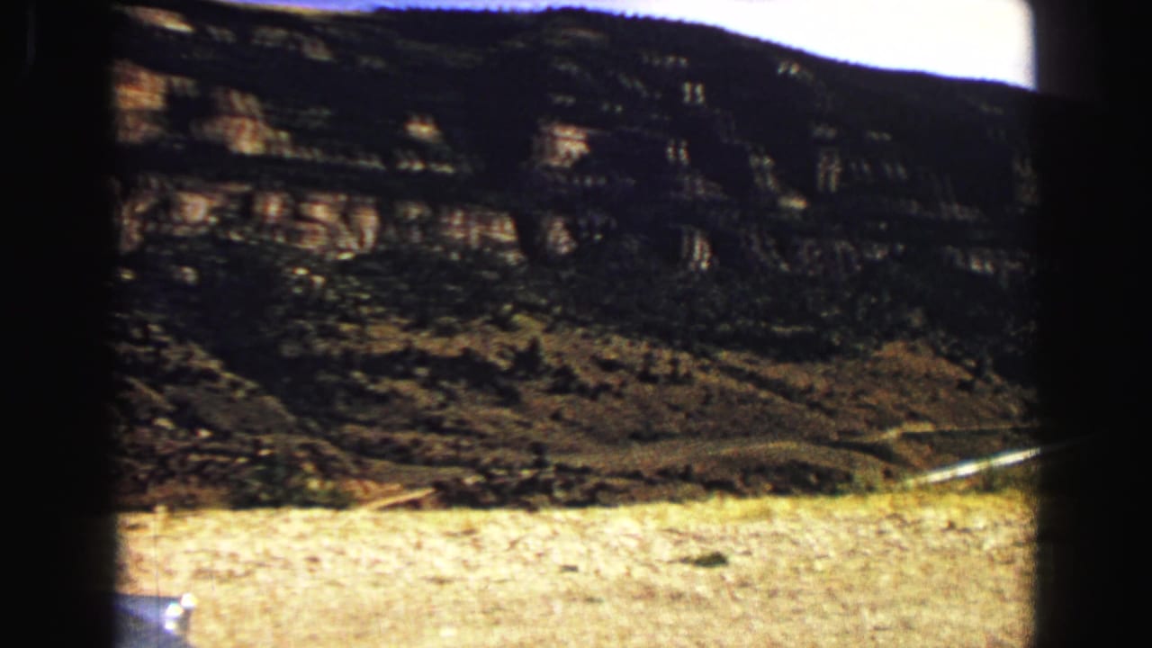 Dark layered hillside with dry roadside ground and a curved road in the distance.
