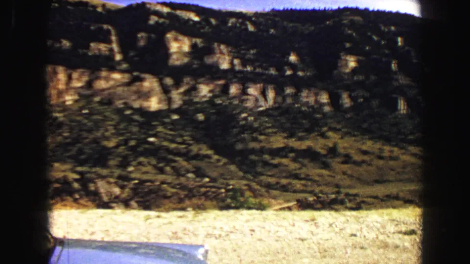 Vintage 8mm view of a dry roadside foreground and dark rocky Yellowstone-area ridge during a 1967 car trip.