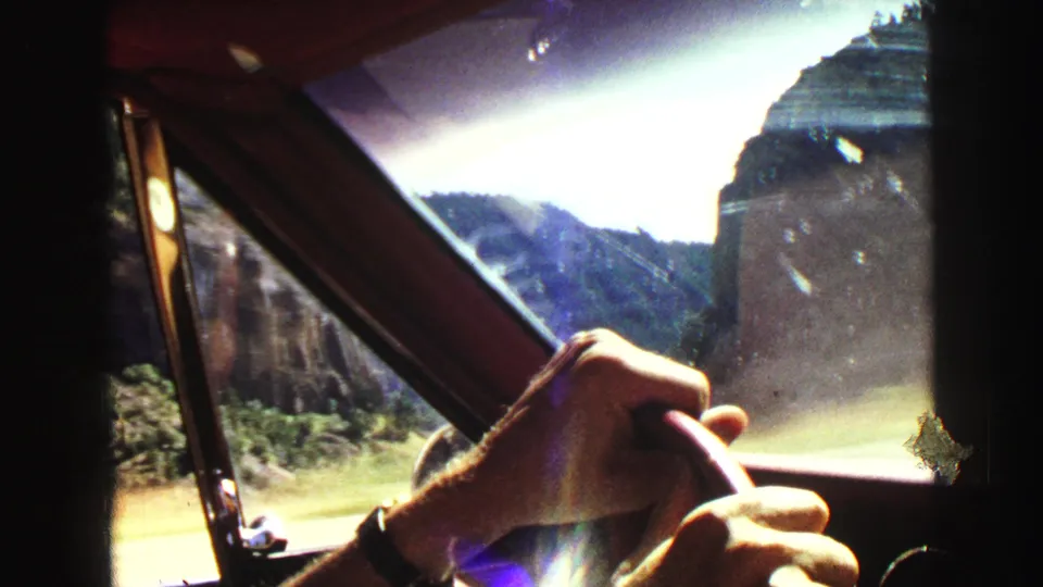 Inside a moving car in 1967 Yellowstone-area footage, a driver's hands hold the wheel as cliffs and sunlit scenery appear ahead.