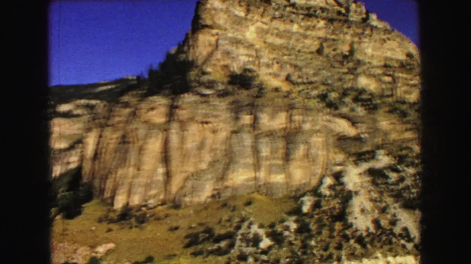 Home-movie view of a large Yellowstone-area cliff with vertical rock textures, scattered evergreens, and dry scrub below.