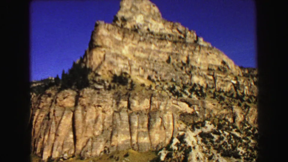 1967 8mm Yellowstone-area frame with a prominent rocky peak, banded cliffs, sparse trees, and deep blue sky.