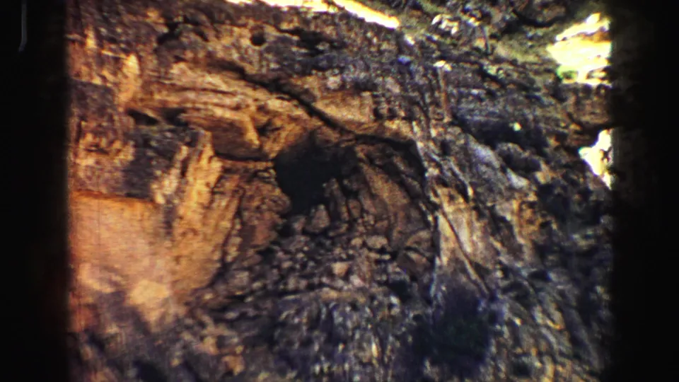 Yellowstone home-movie frame showing a closer rugged rock formation with warm tan stone, dark fissures and scattered vegetation.