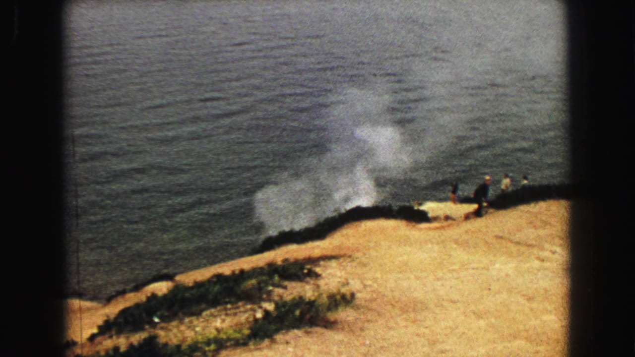 Steam rises from a hillside above a dark lake with small distant visitors nearby.