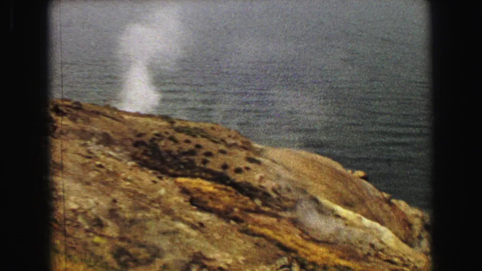 1967 Yellowstone 8mm frame showing a taller white steam plume from a barren hillside overlooking dark water.