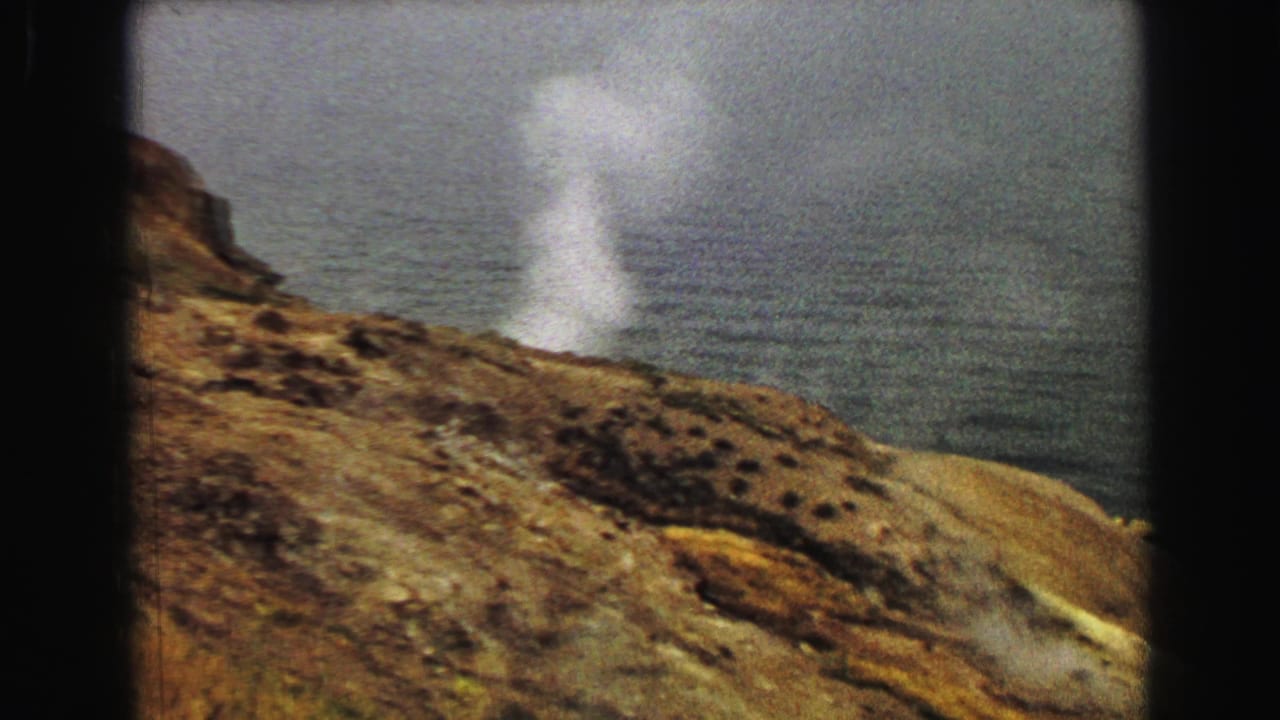 White steam plume rising from a rocky hillside above a broad body of water.