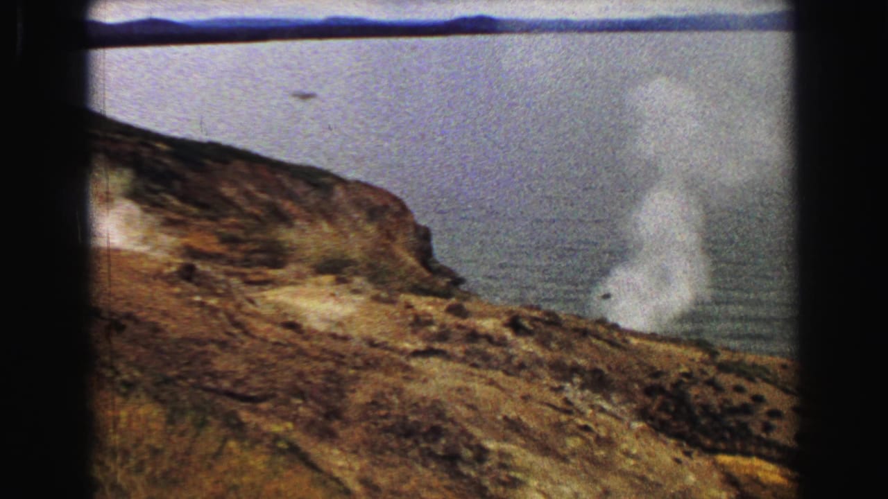 Rocky slope with white steam near water and a distant shoreline.