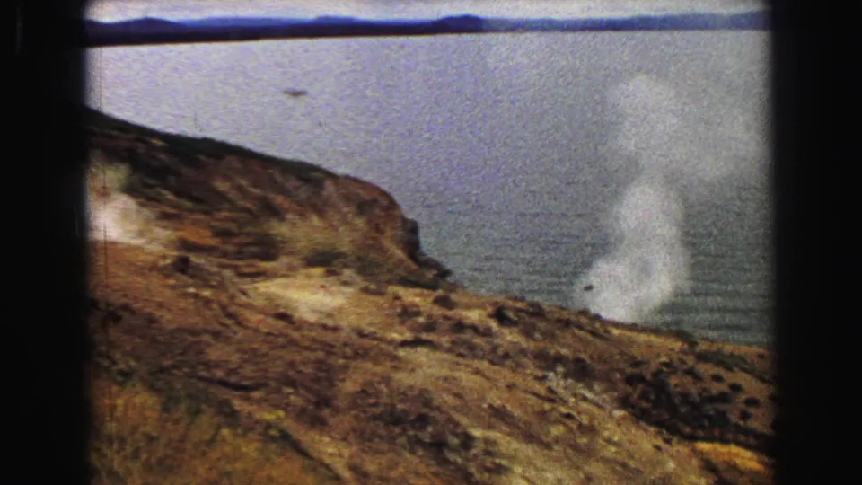 1967 Yellowstone home-movie frame showing a geothermal steam plume on a brown hillside beside a calm lake or river.