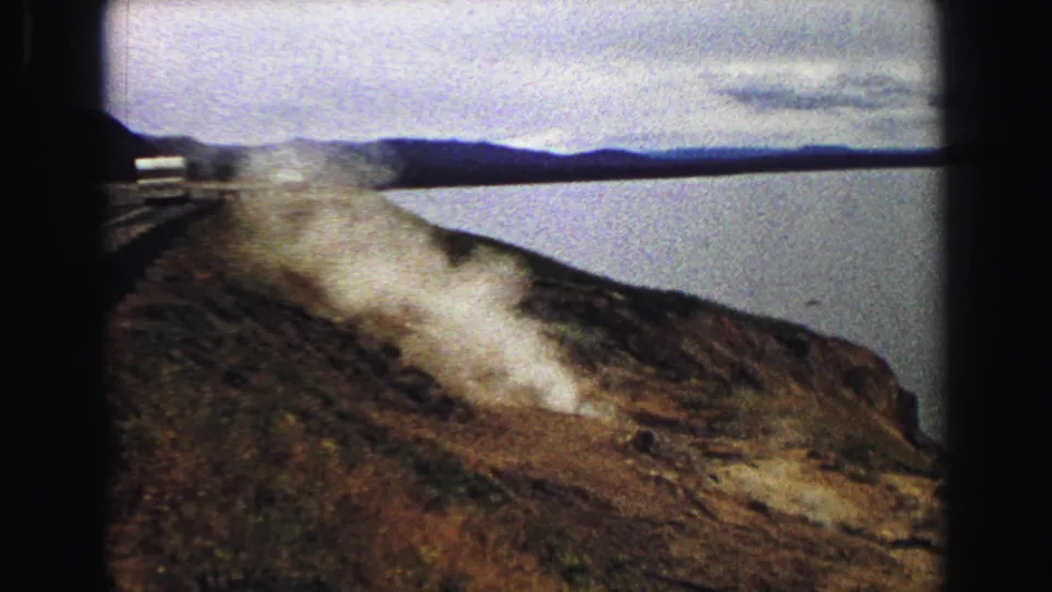 1967 Yellowstone home-movie still showing steam over a rugged lakeside slope, framed by dark 8mm film edges.