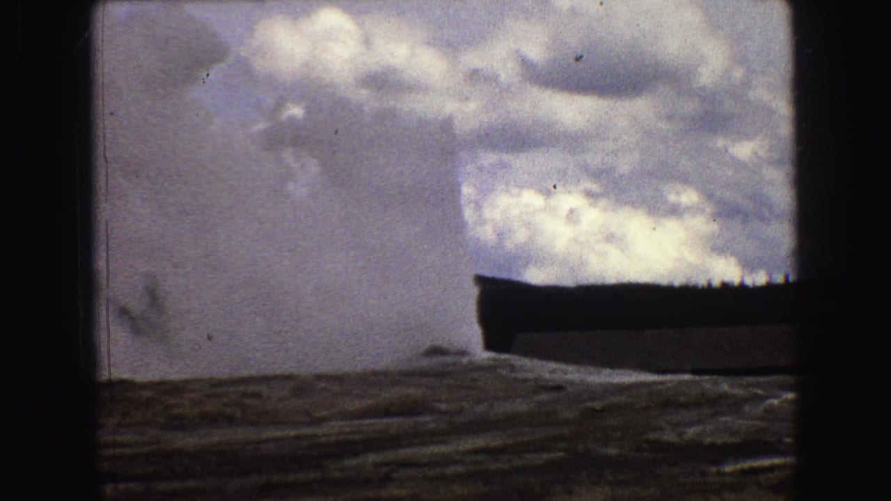 Geyser plume rising behind rocky ground under a cloudy sky.