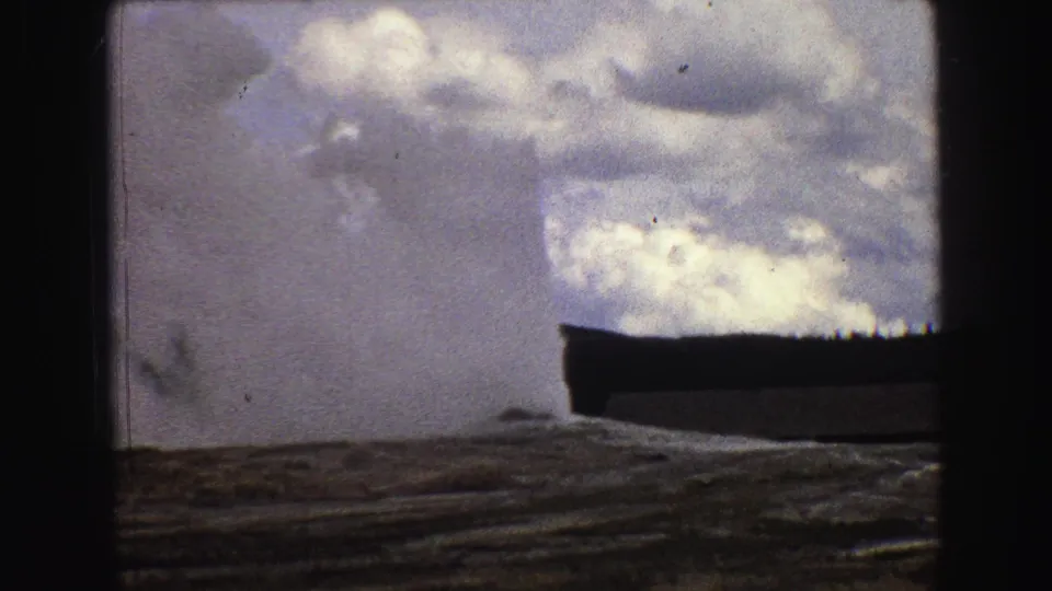 Yellowstone geyser eruption seen from a distance on 1967 8mm film, with cloudy sky and dark film gate edges.
