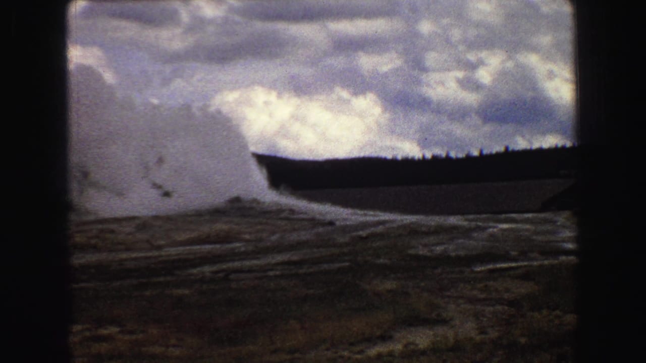 Geyser steam plume beside a dark ridge and cloudy sky.