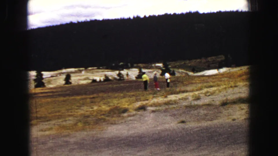 A small group of Yellowstone visitors stands on open terrain near a geyser basin, with a forested ridge in the background.