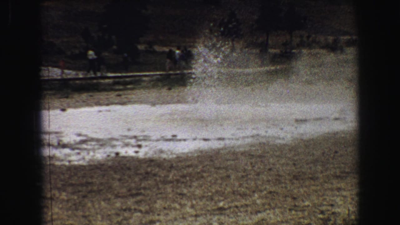 Small geyser spurting water from pale ground with visitors in the distance.