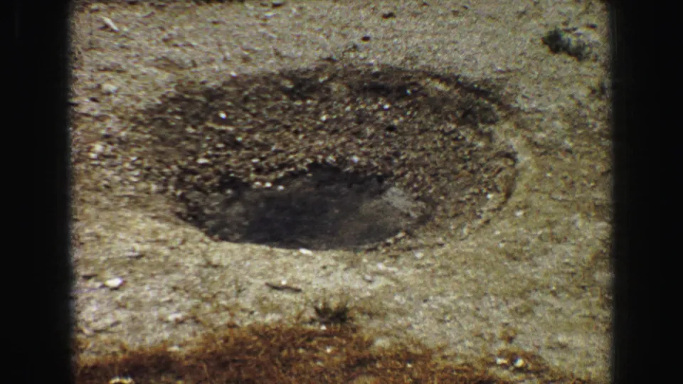 1967 8mm Yellowstone view of a dark geyser opening in stony mineral ground with distant visitors beyond the spray area.