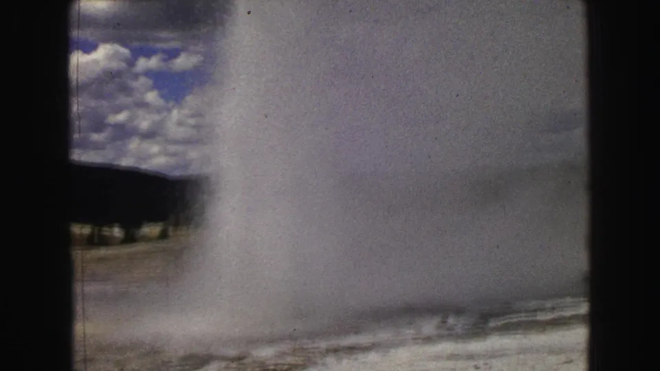 1967 Yellowstone home-movie frame showing Old Faithful erupting, with steam and water rising against clouds and a dark treeline.