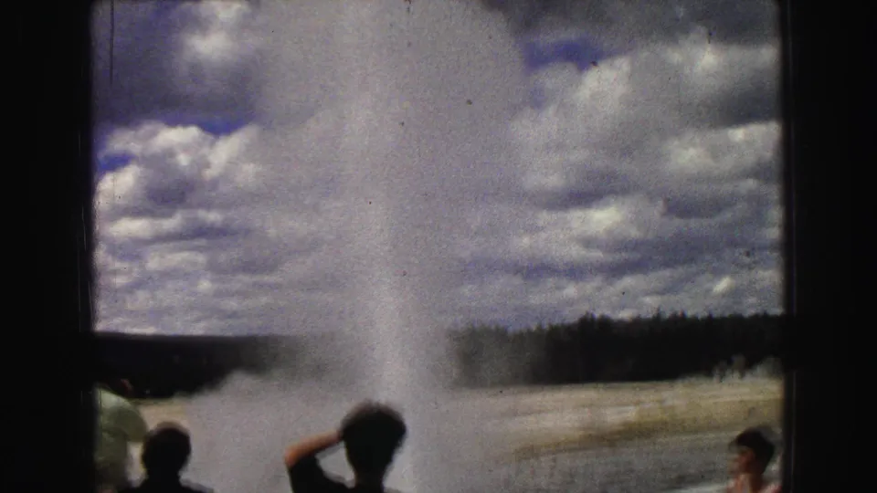 1967 Yellowstone frame with visitors in silhouette watching Old Faithful erupt beneath a dramatic clouded sky.