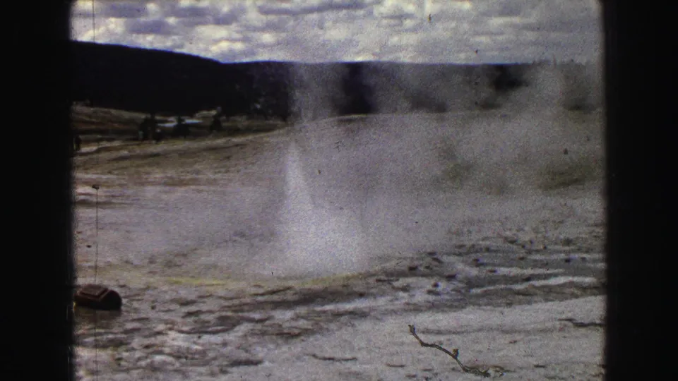 1967 Yellowstone 8mm frame showing a geyser eruption with steam over mineral-streaked ground, dark hills, and tiny distant visitors or vehicles.