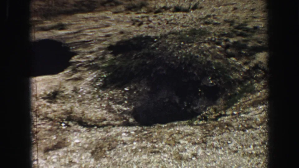 1967 Yellowstone 8mm close view of a dark geothermal vent or pool in mineral-streaked ground, with grain, soft focus, and frame shadowing.
