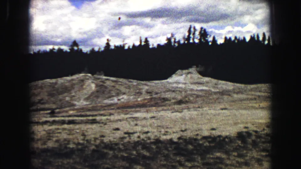 1967 8mm Yellowstone wide shot of pale geyser terrain beneath a dark tree line and cloudy sky.