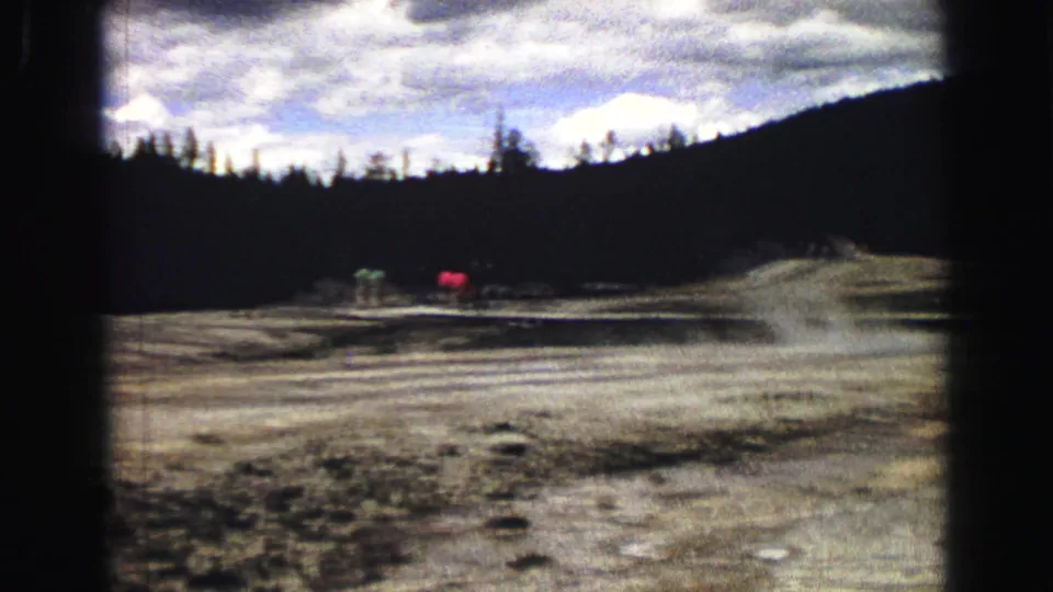 1967 Yellowstone home-movie view across the Old Faithful basin, with distant tourists, steam, dark trees, and cloudy sky.