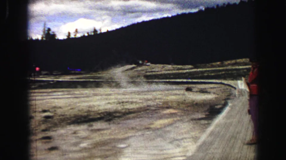 1967 Yellowstone frame showing the Old Faithful viewing boardwalk, pale thermal ground, distant visitors, and one person at the right edge.