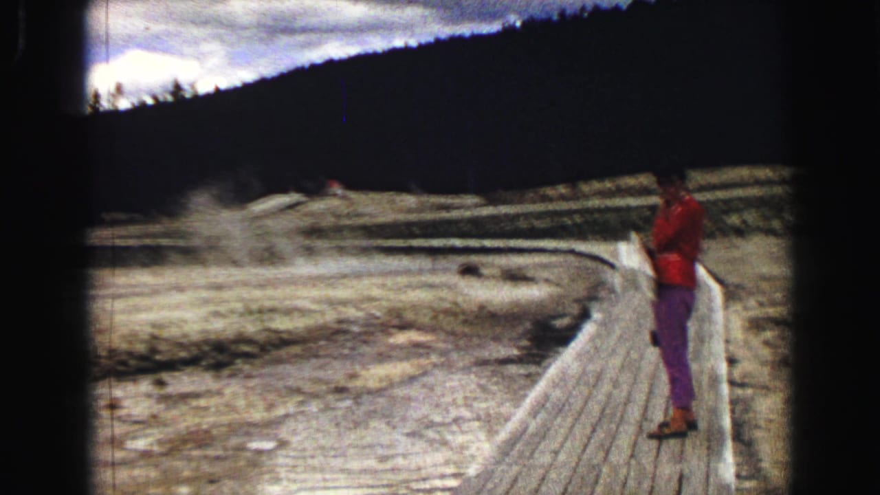 Visitor in red jacket standing on a boardwalk near steaming geothermal ground.