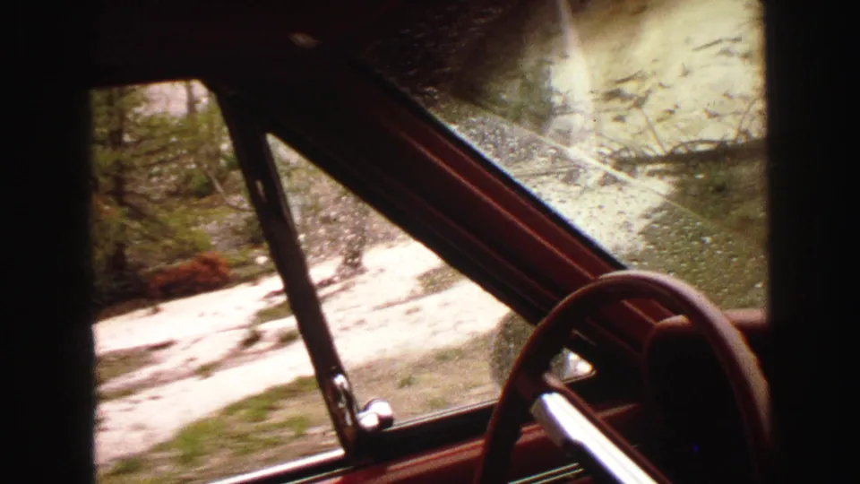Final 1967 travel frame from inside the vehicle, showing steering wheel, rain-speckled window, and road surface outside.