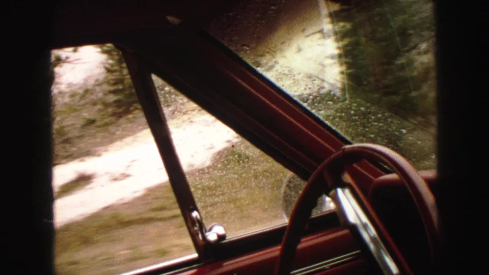 1967 Yellowstone-area home-movie frame from inside a car, looking past a red door frame and wet glass toward a blurred dirt road.