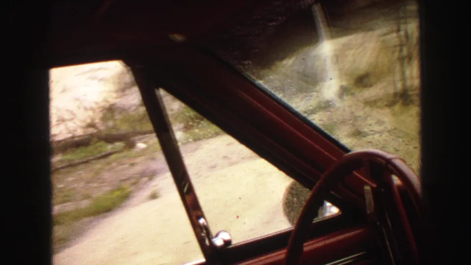 Inside-car view in the 1967 Yellowstone clip, with rainy glass, red trim, steering wheel, and rocky roadside passing outside.